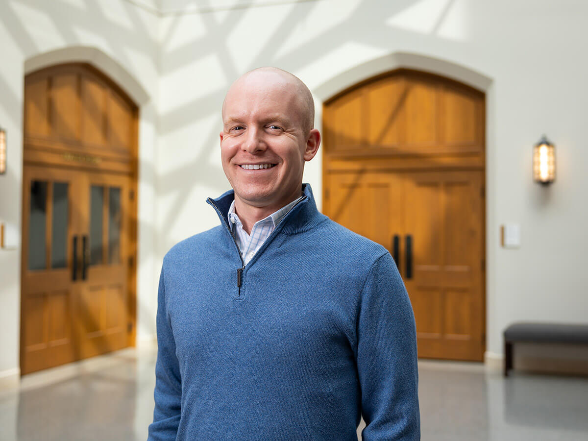 Smiling bald man in a blue quarter-zip sweater over a plaid shirt, standing in a brightly lit hallway with large wooden arched doors.