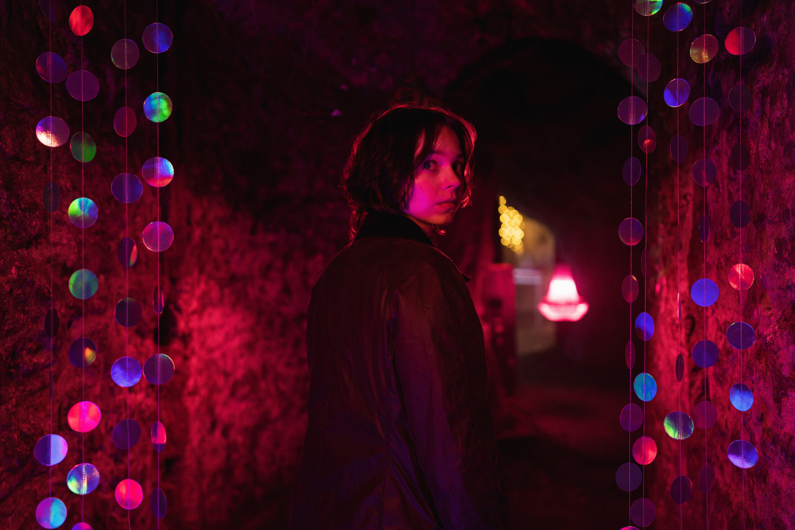 "Person standing in a dimly lit tunnel with exposed stone walls, illuminated by vibrant pink, purple, and blue lights and surrounded by hanging colorful circles, creating a mysterious and moody atmosphere."