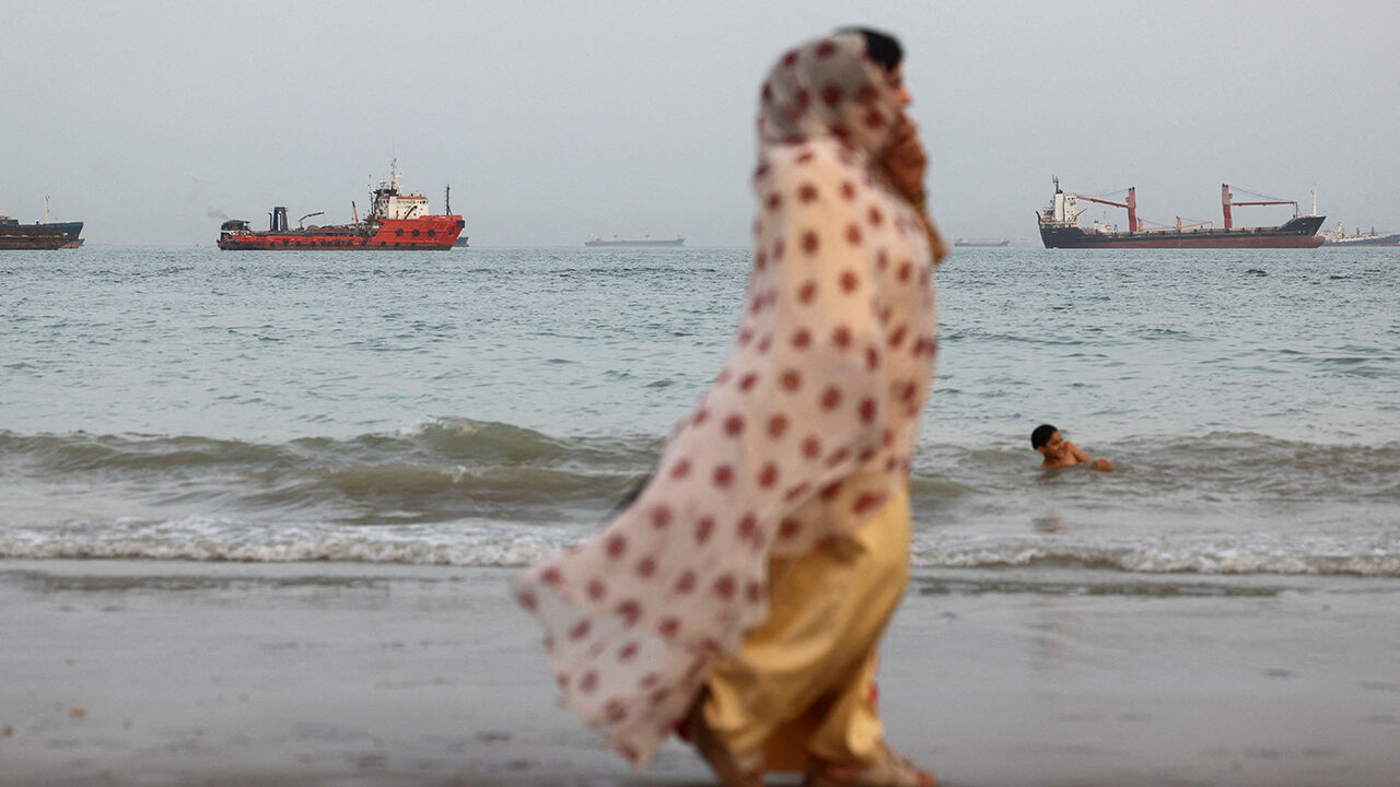 A woman walks on the beach while a boy swims, with vessels in the Strait of Hormuz visible near Bandar Abbas, Iran, May 22nd 2026
