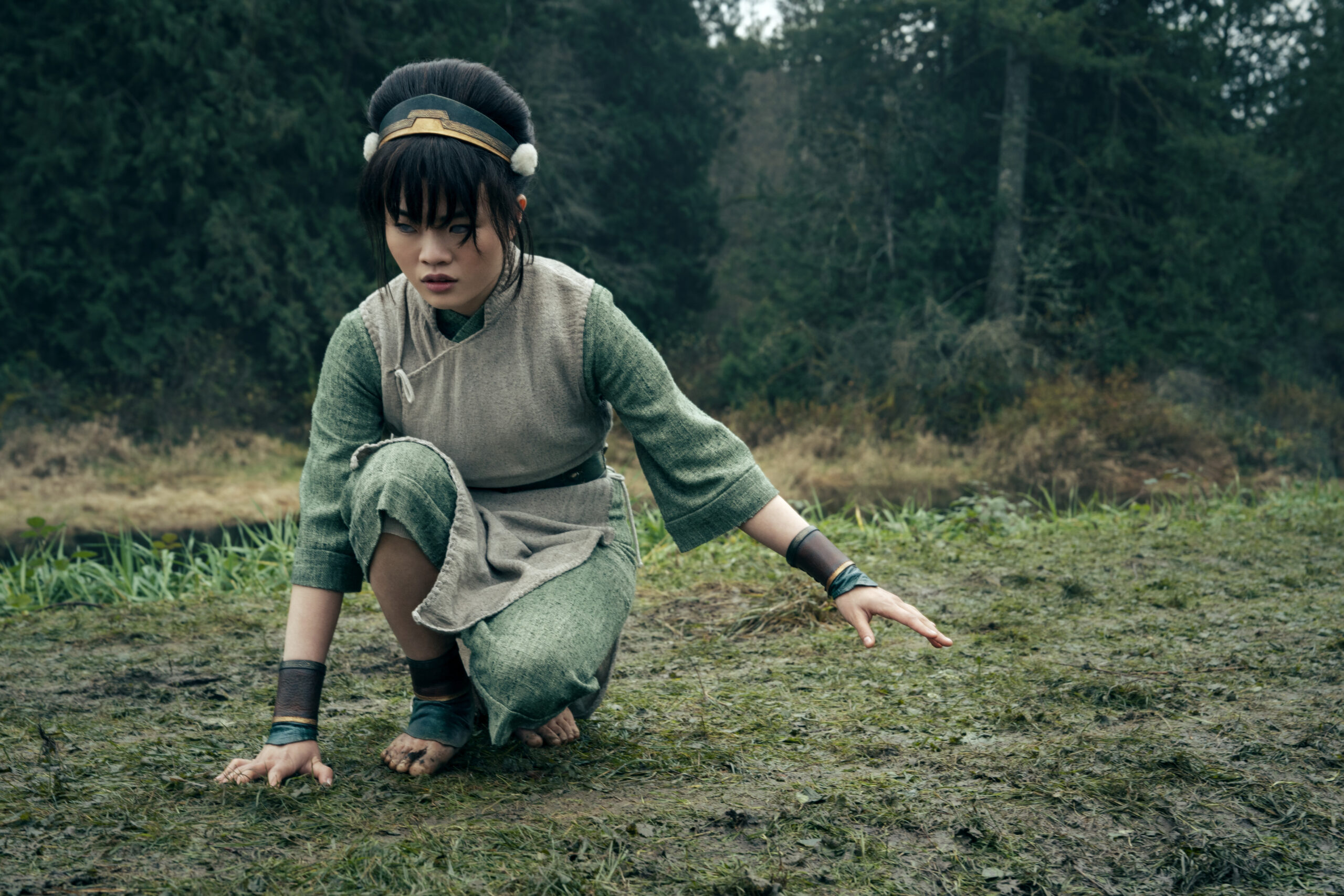 "A young girl with black hair wearing green clothes crouches down in the woods with bare feet on grass and right hand touching the ground. "