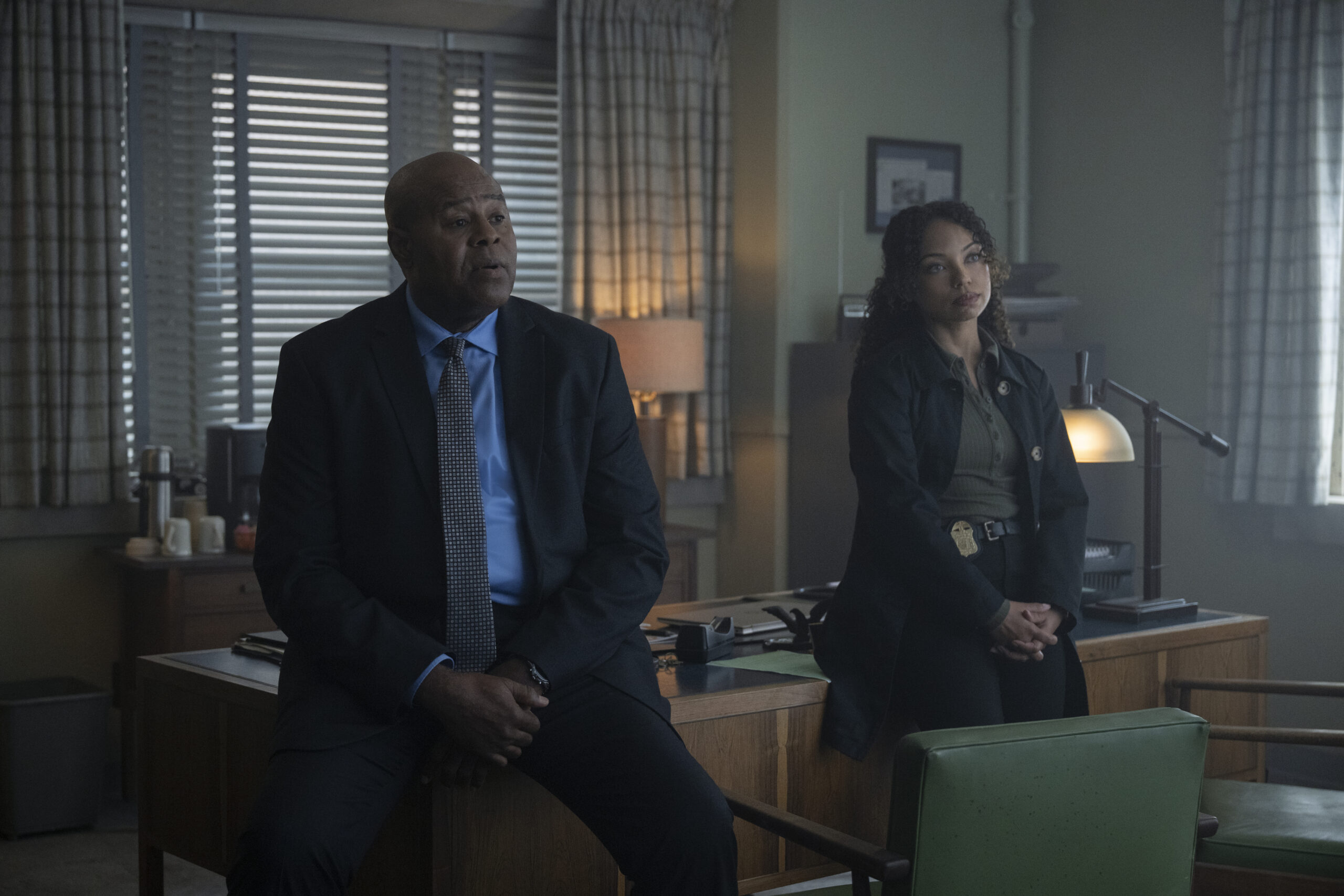 "Two detectives sit and lean against a wooden desk in a dimly lit office with large windows, blinds, lamps, and paperwork, suggesting a serious conversation or investigation."