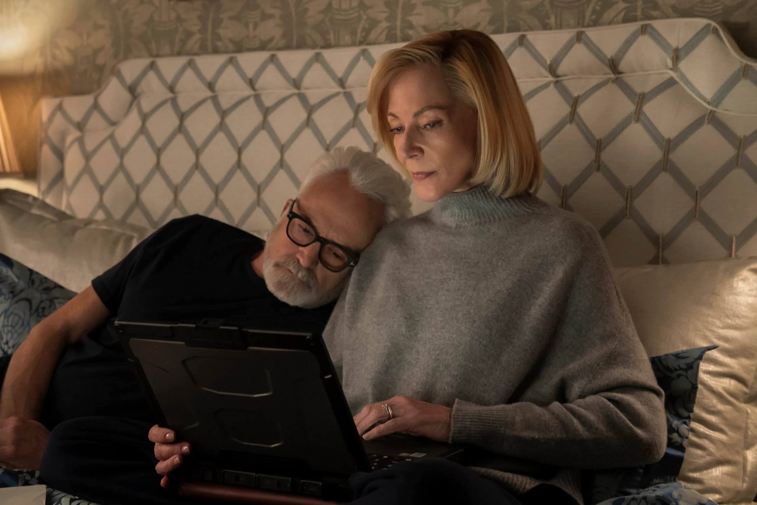 "Older couple relaxing in bed together, man resting his head on woman's shoulder while she uses a tablet, cozy bedroom setting with patterned headboard and soft lighting."