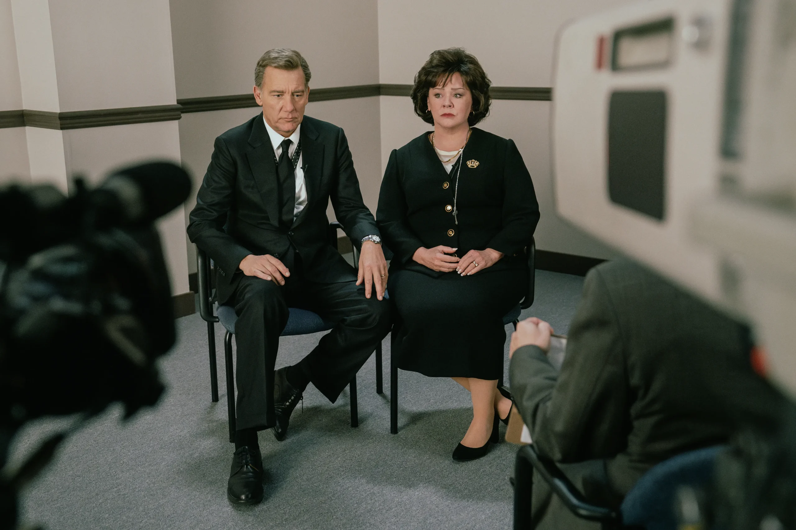 "Two people in formal attire sit side by side in a plain office-like room, holding hands, being filmed by a video camera during an interview or press conference."