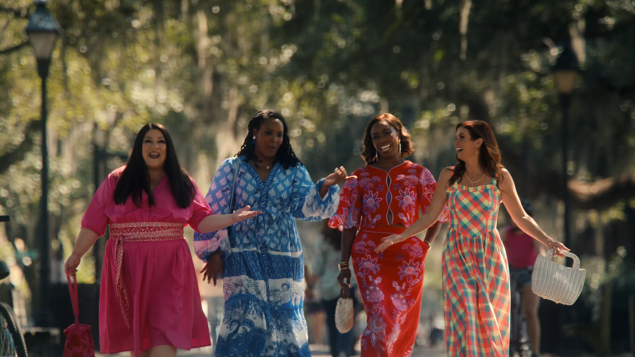 "Four women in colorful dresses walk together outdoors along a sunlit, tree-lined path, smiling and talking, with relaxed park surroundings and dappled sunlight in the background."