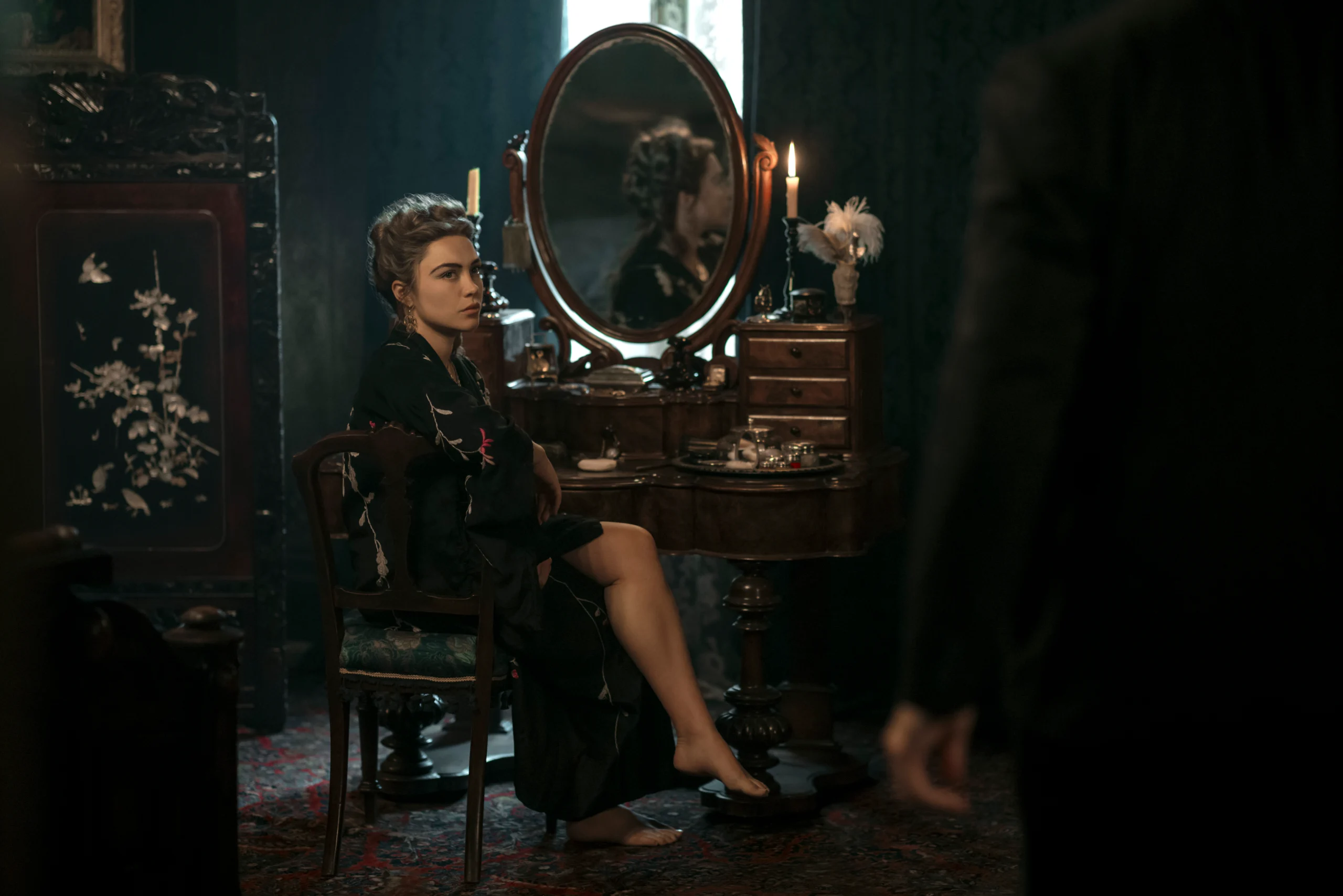 "Woman in dark robe sits at vintage vanity with mirror and candles in dimly lit, elegant room; another person stands in foreground, partly visible."